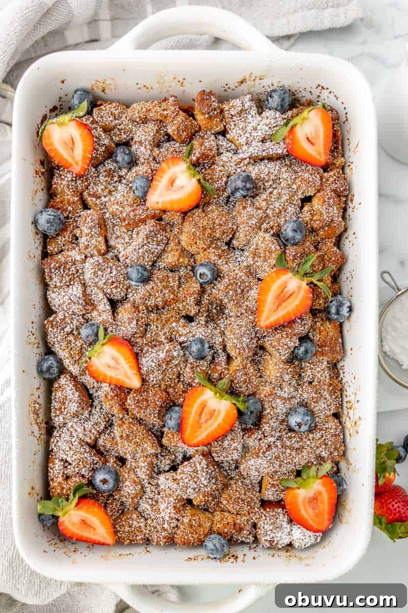 Overnight Brioche French Toast Casserole Recipe 11 Baked dish topped with powdered sugar, halved strawberries, and blueberries in a rectangular white baking dish on a white surface.