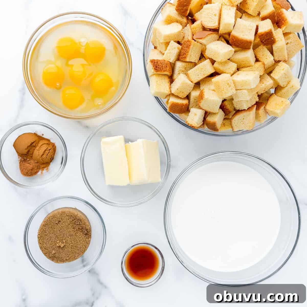 Overnight Brioche French Toast Casserole Recipe 3 Bowls with bread cubes, eggs, cinnamon, nutmeg, butter, milk, brown sugar, and vanilla on a marble surface.