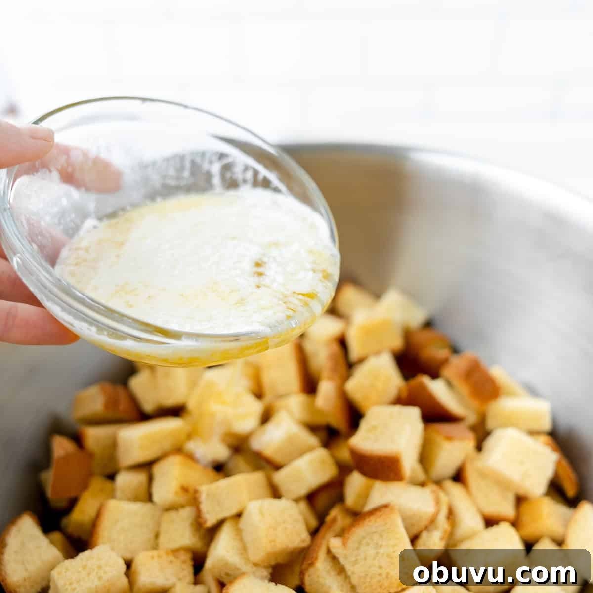 Overnight Brioche French Toast Casserole Recipe 4 Hand pouring melted butter over cubed bread in a metal bowl.