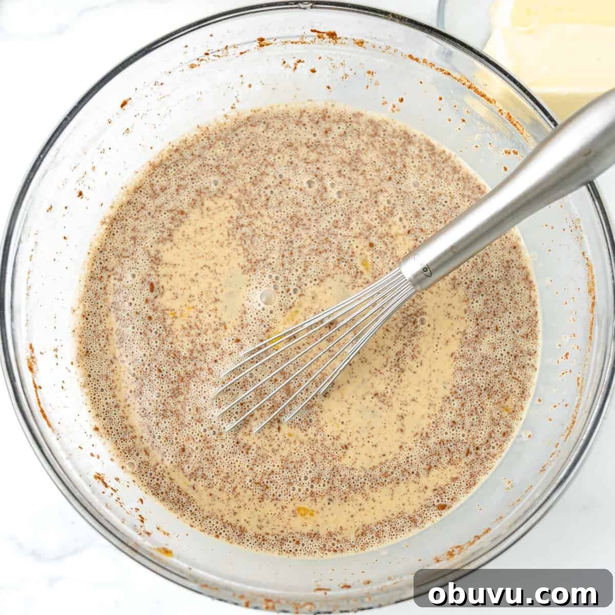 Overnight Brioche French Toast Casserole Recipe 5 A metal whisk is in a glass bowl containing a brown, speckled liquid mixture. A block of butter is visible in the background.