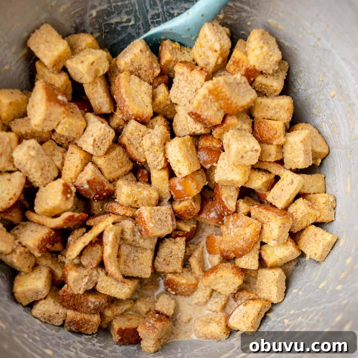 Overnight Brioche French Toast Casserole Recipe 6 Cubed bread pieces coated in a mixture of spices and liquid sit in a mixing bowl with a blue spatula.