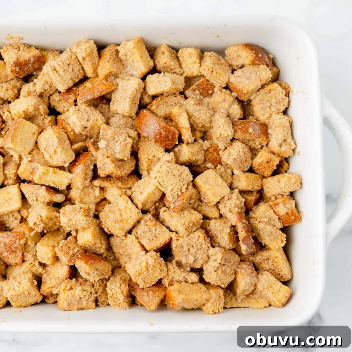 Overnight Brioche French Toast Casserole Recipe 7 A white baking dish filled with cubed bread coated in a mixture, likely for stuffing or bread pudding, on a marble surface.