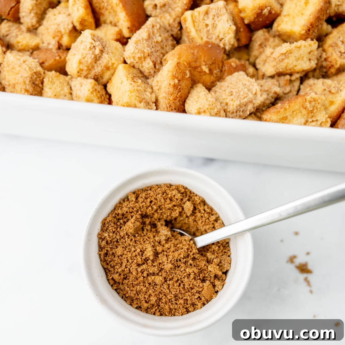 Overnight Brioche French Toast Casserole Recipe 8 White baking dish filled with cubed bread pieces, beside a small bowl of brown sugar with a spoon on a marble surface.