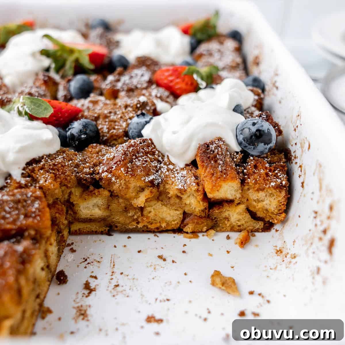 Overnight Brioche French Toast Casserole Recipe 10 Baked French toast casserole with missing piece, garnished with blueberries, strawberries, and dollops of whipped cream in a white dish.