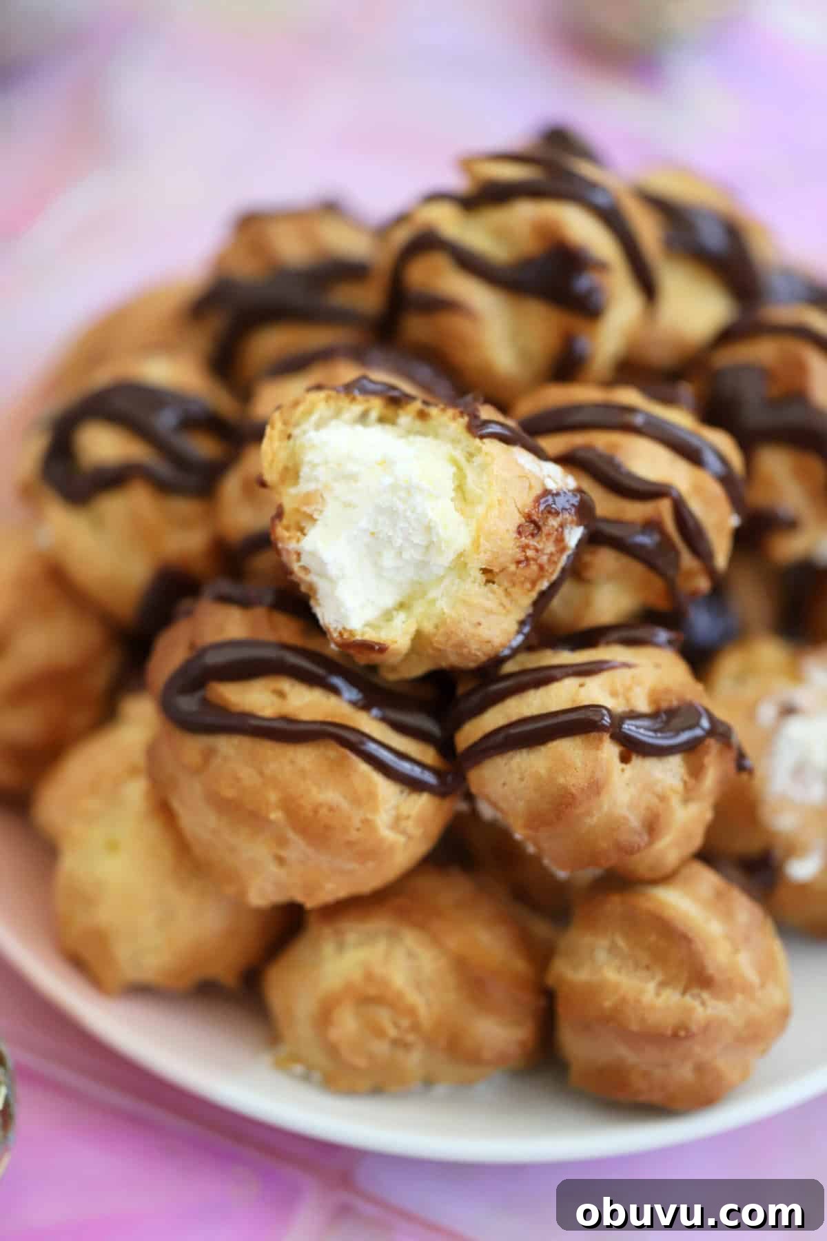 A plate of gluten free profiteroles drizzled with chocolate sauce, with one cut open to show the whipped cream filling.