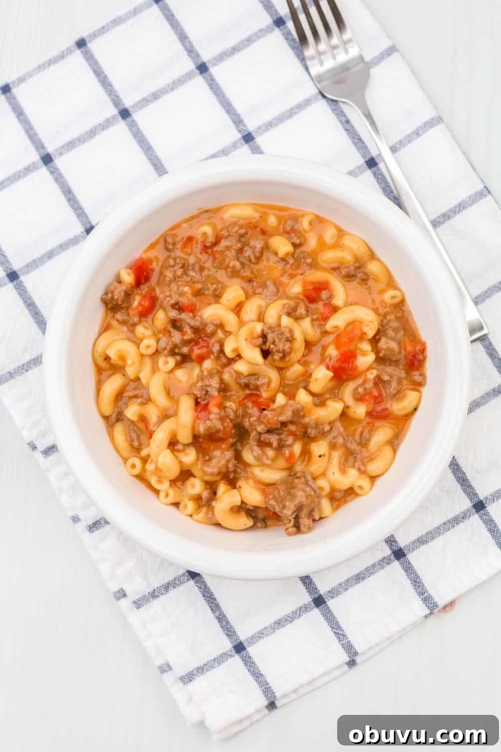 a white bowl of pressure cooker cheeseburger macaroni on top of a blue and white checkered napkin, with a fork next to it.