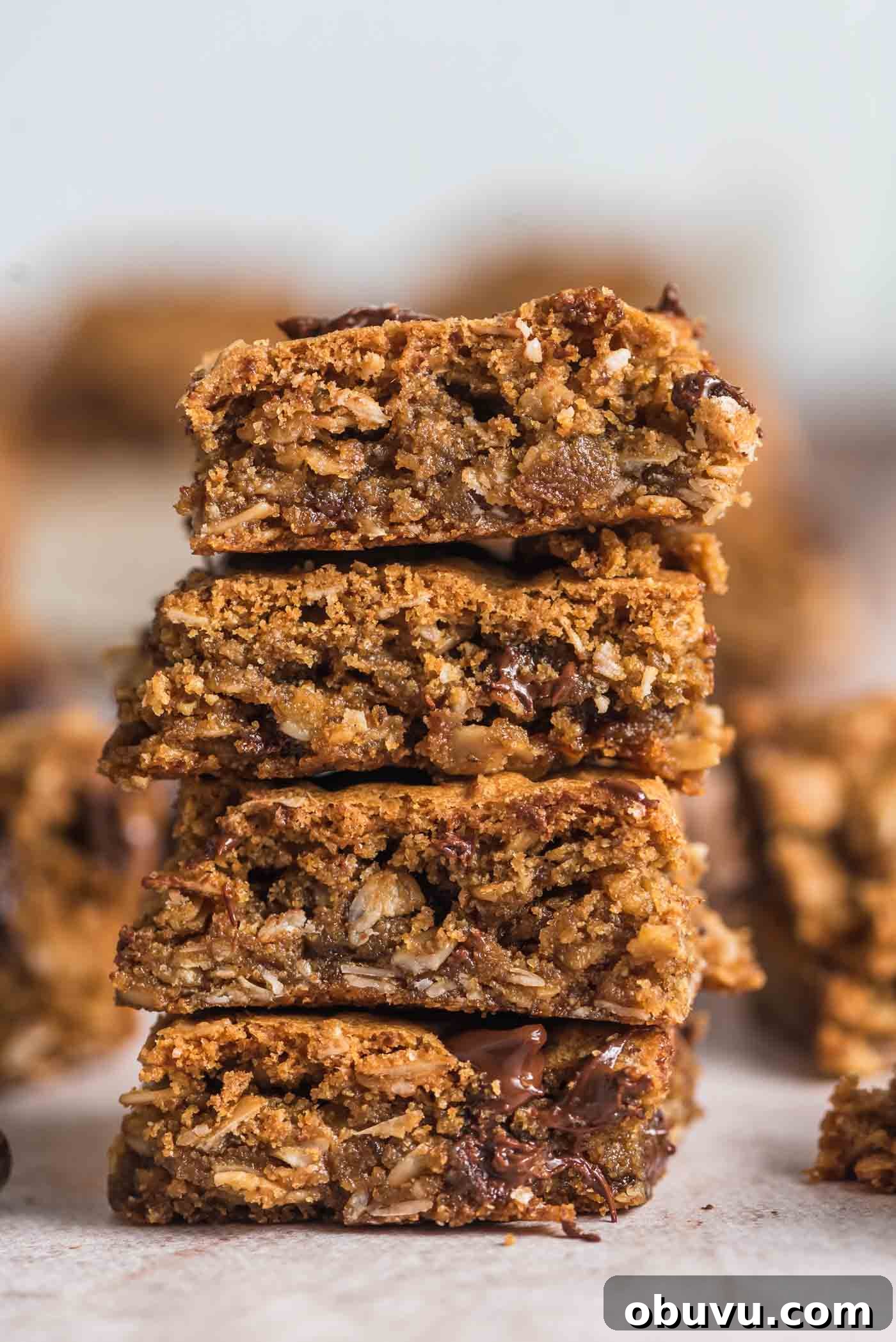 A stack of 4 vegan and gluten-free oatmeal cookie bars with chocolate chips in them. More bars can be seen in the background.