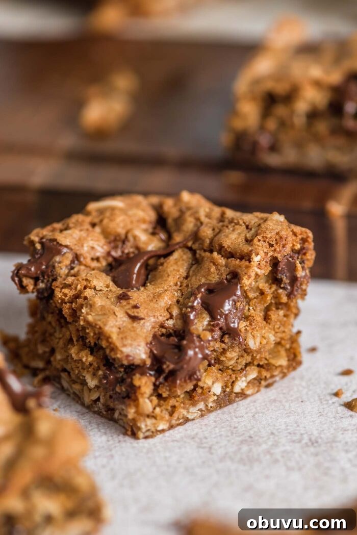 An oatmeal cookie bar sitting on a counter with a cutting board behind it with more bars on it. You can see the texture and melted chocolate chips inside the bar.